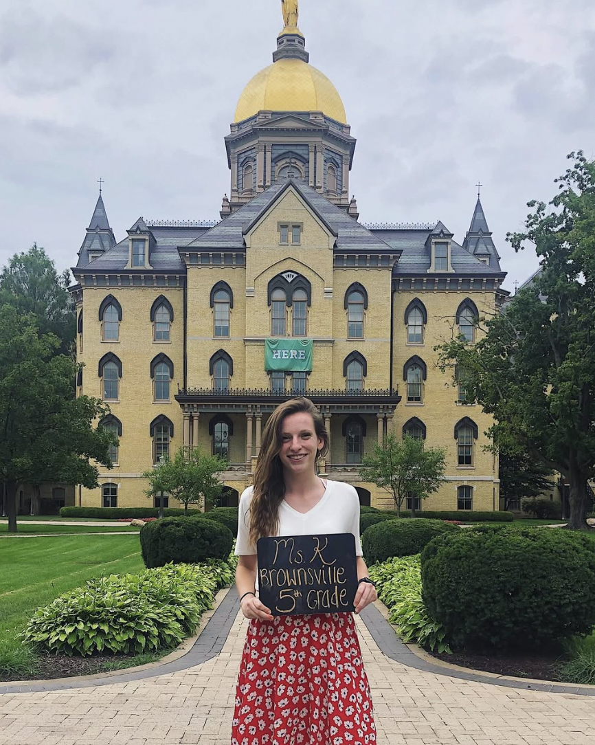 brunette standing in front of the Notre Dame golden dome holding a sign saying "Ms. K Brownsville 5th Grade"